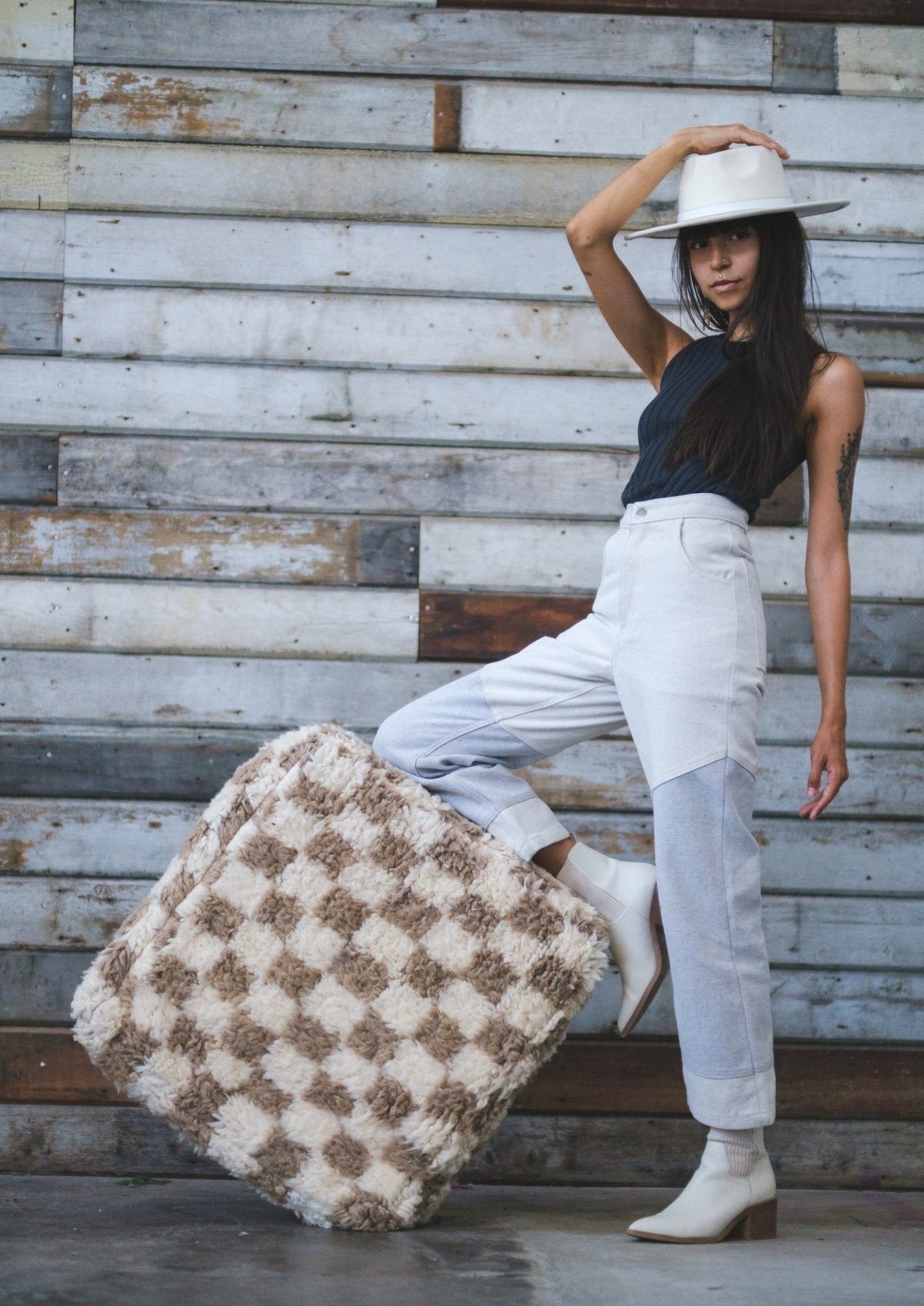 hot girl with long brown hair holds her white hat with one hand while leaning on a moroccan floor cushion checkered white and tan. She is wearing soluna collective top and pants organic natural fibers with a rustic wood wall backdrop
