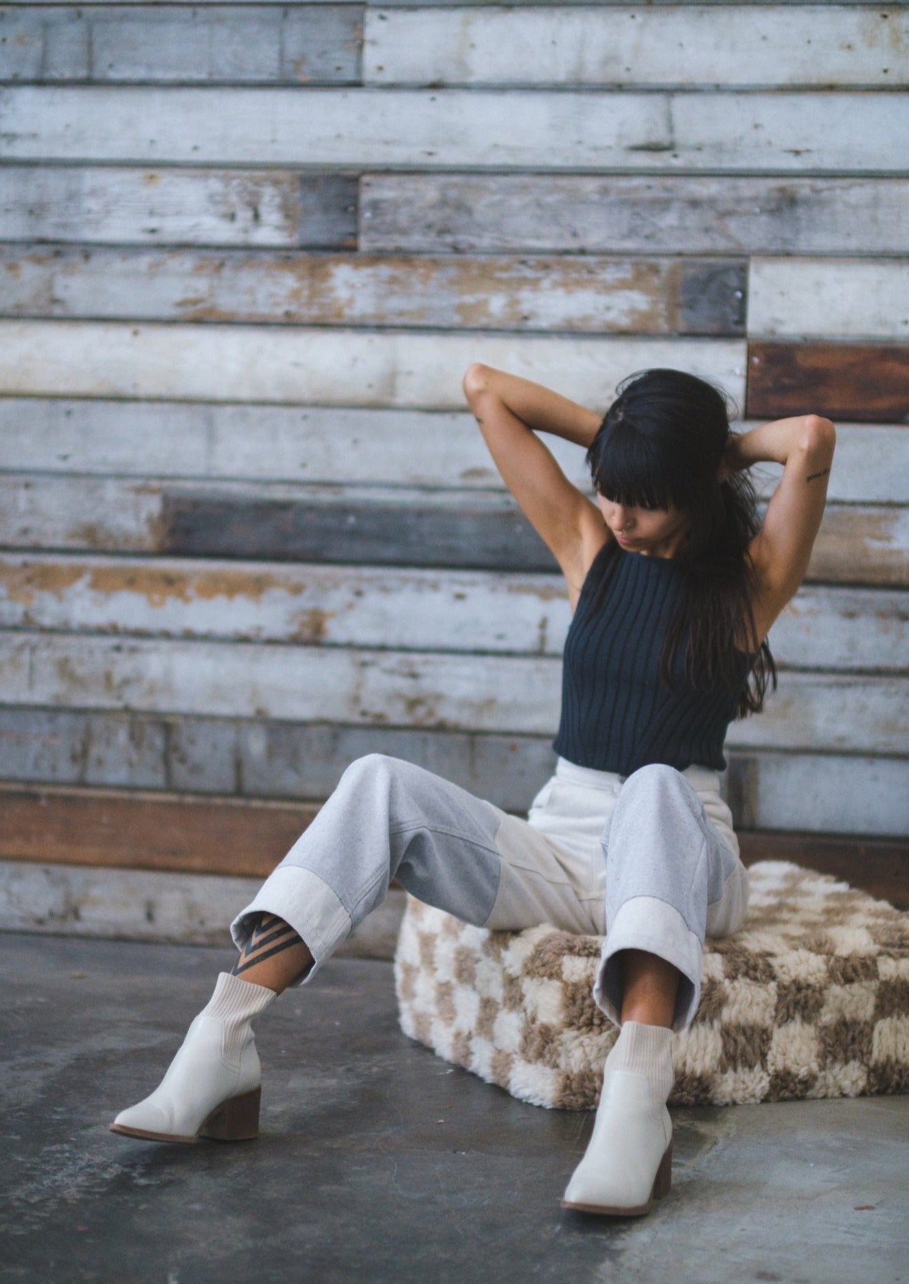 gorgeous girl with long black hair ties it back while she sits on a moroccan floor cushion checkered. She is wearing soluna collective top and bottom made from organic fibers designed in portland oregon. location is cement floors with rustic wood wall