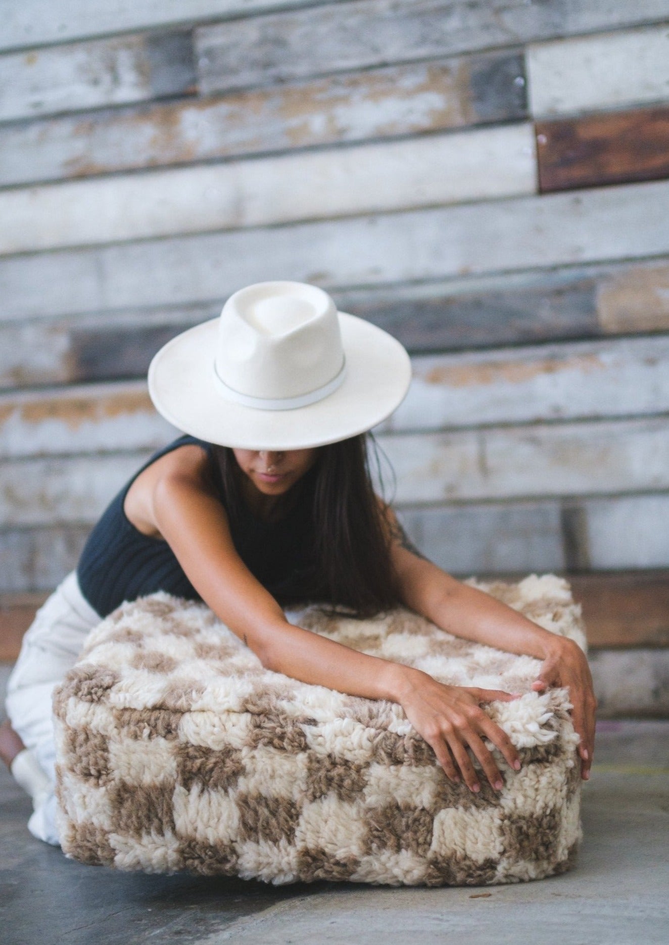 gorgeous and mysterious girl wearing white hat holds and hugs a moroccan floor cushion thats checkered white and tan in a rustic location. 100% wool.