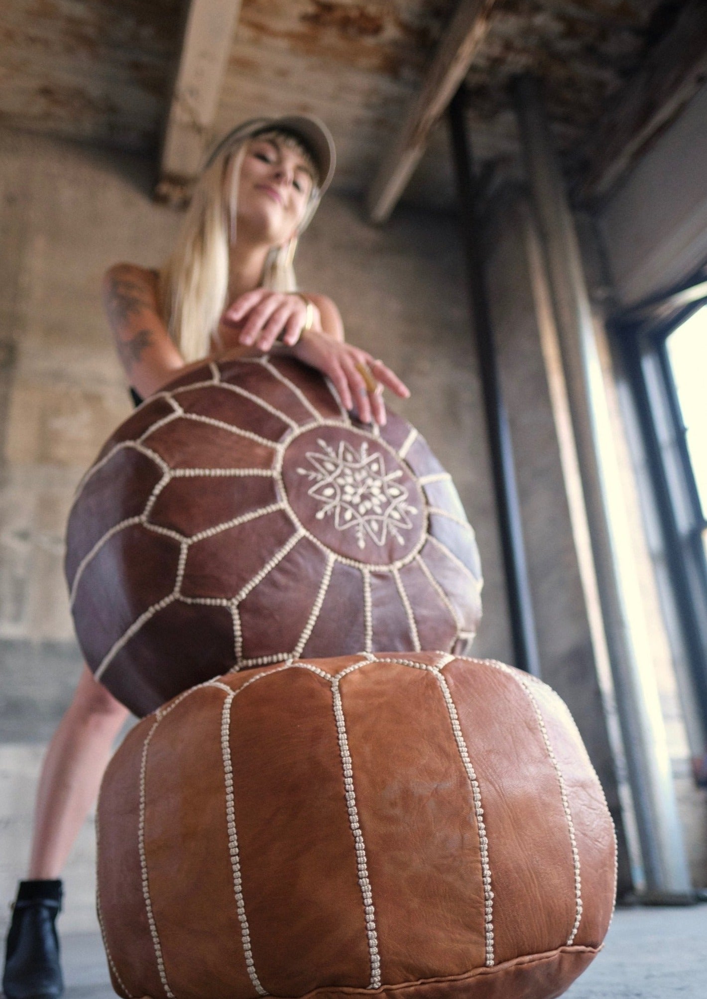 girl stands proudly in her loft apartment, light shining through the window highlighting her glowing skin and leather poufs she leans on with relaxed hands