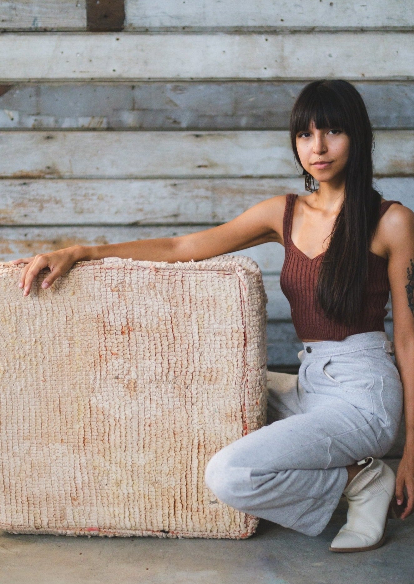 Moroccan floor cushion is on ground in rustic space with refurbished wood. girl rests her arm on it and smiles at camera. 