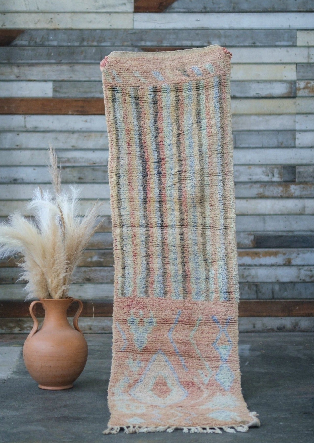 simple earth tones woven with wool on this moroccan runner displayed against a barn wall with rustic wood and a ceramic vase with sea grass. 