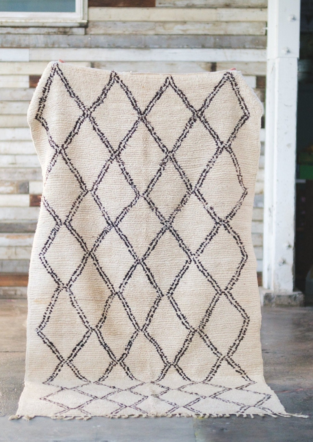 classic beni ourain rug is displayed against a rustic barn wall and cement floors. white wool decorated with black diamonds all natural dye