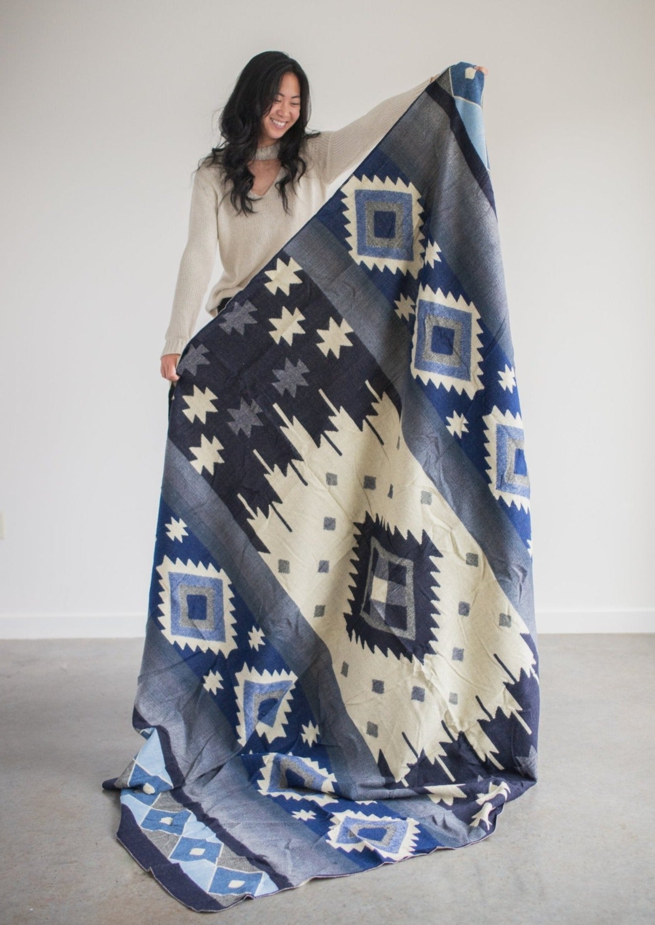 asian girl wearing comfy sweater holds up an alpaca blanket made in Ecuador so you can see the full design.