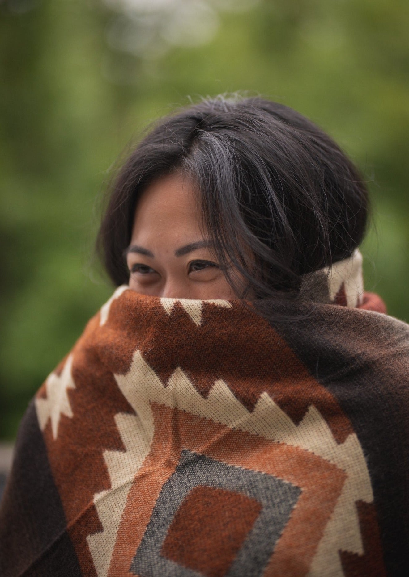 asian girl cozies up with her soft alpaca blanket in the fall outside. Her eyes are peeking out from behind the brown blanket