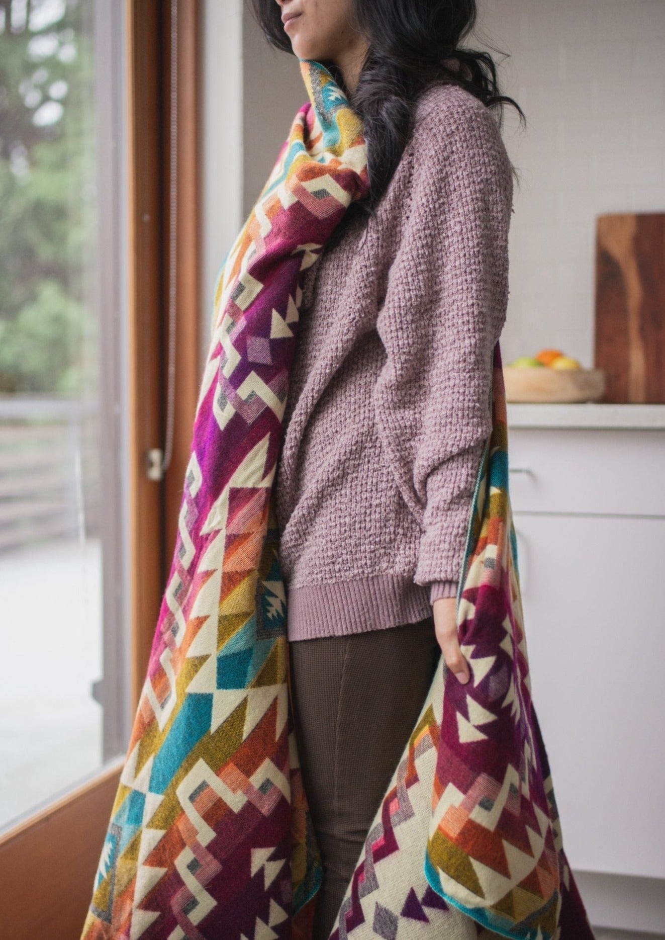 Asian girl wearing cozy clothes in her kitchen looking out the window with her alpaca blanket draped around her for warmth