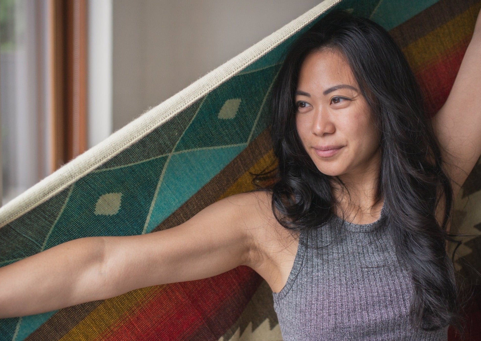 Asian girl stands with arms wide open holding her cozy alpaca blanket behind her looking out the window. Her hair is long and curled. 