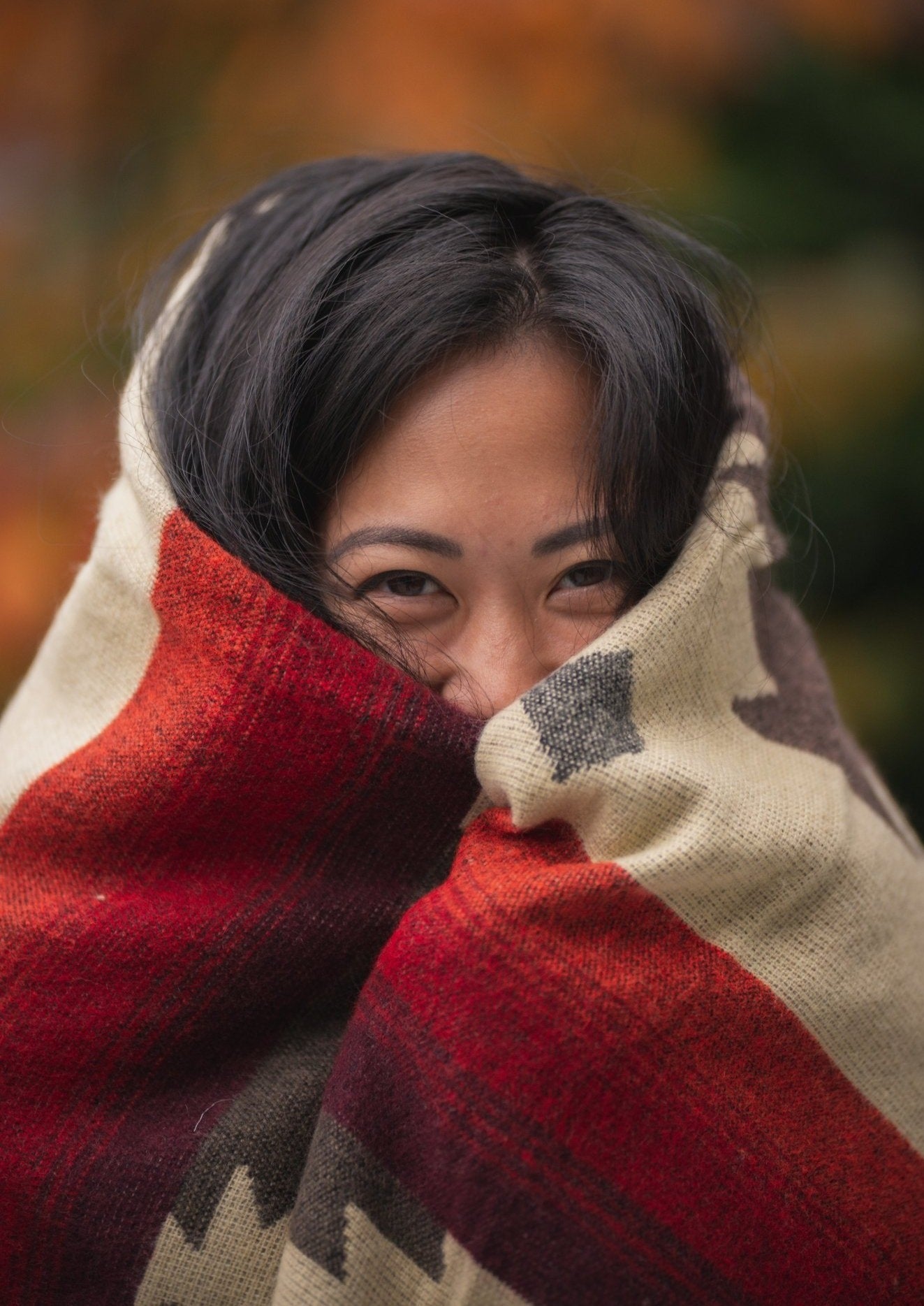 Asian girl wraps up in a cozy alpaca blanket outside during fall. The blanket keeps her face warm and her eyes are smiling.