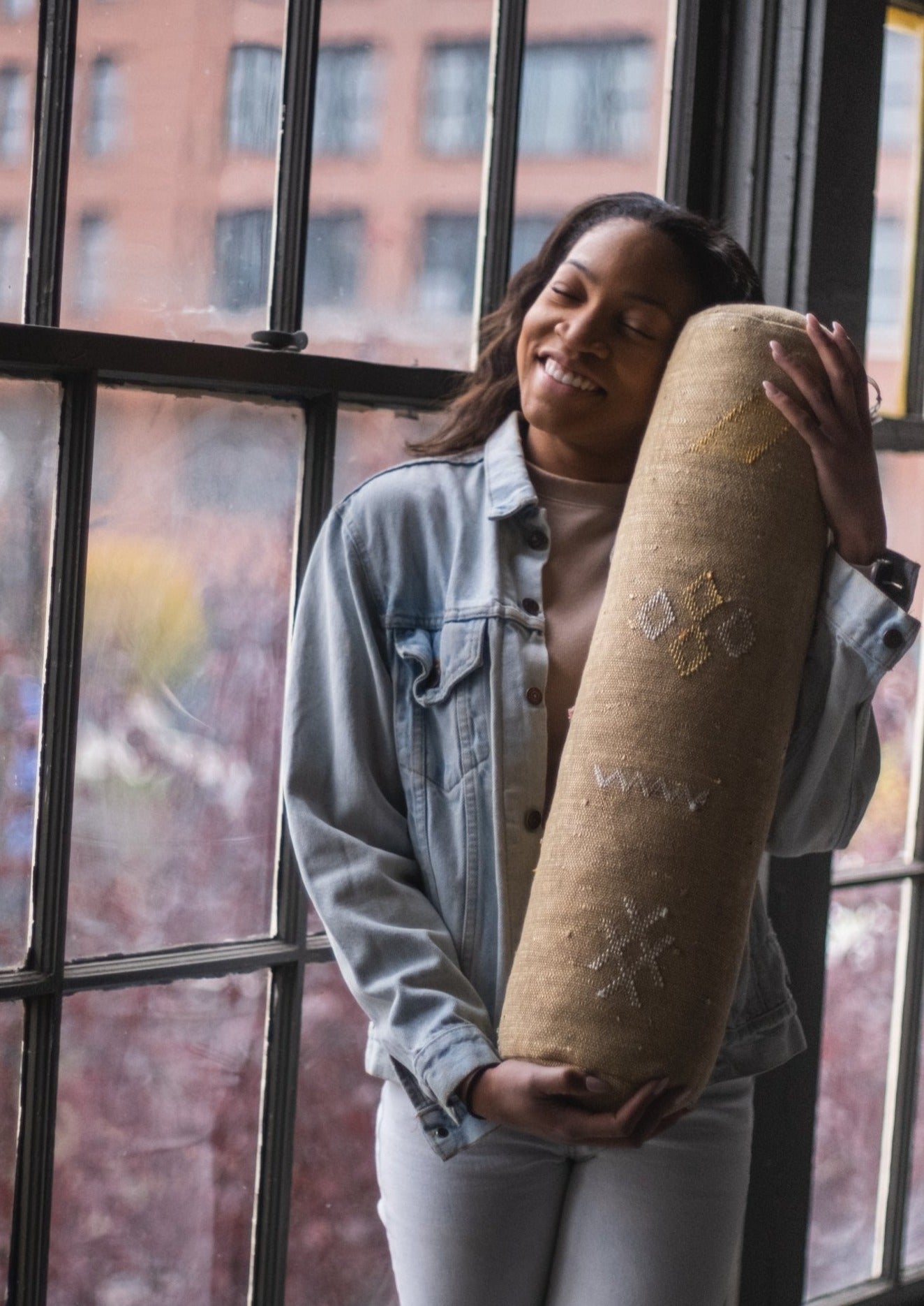 African American young girl stands by window in loft apartment holding her favorite pillow made from vegan friendly silk fibers in Morocco. 
