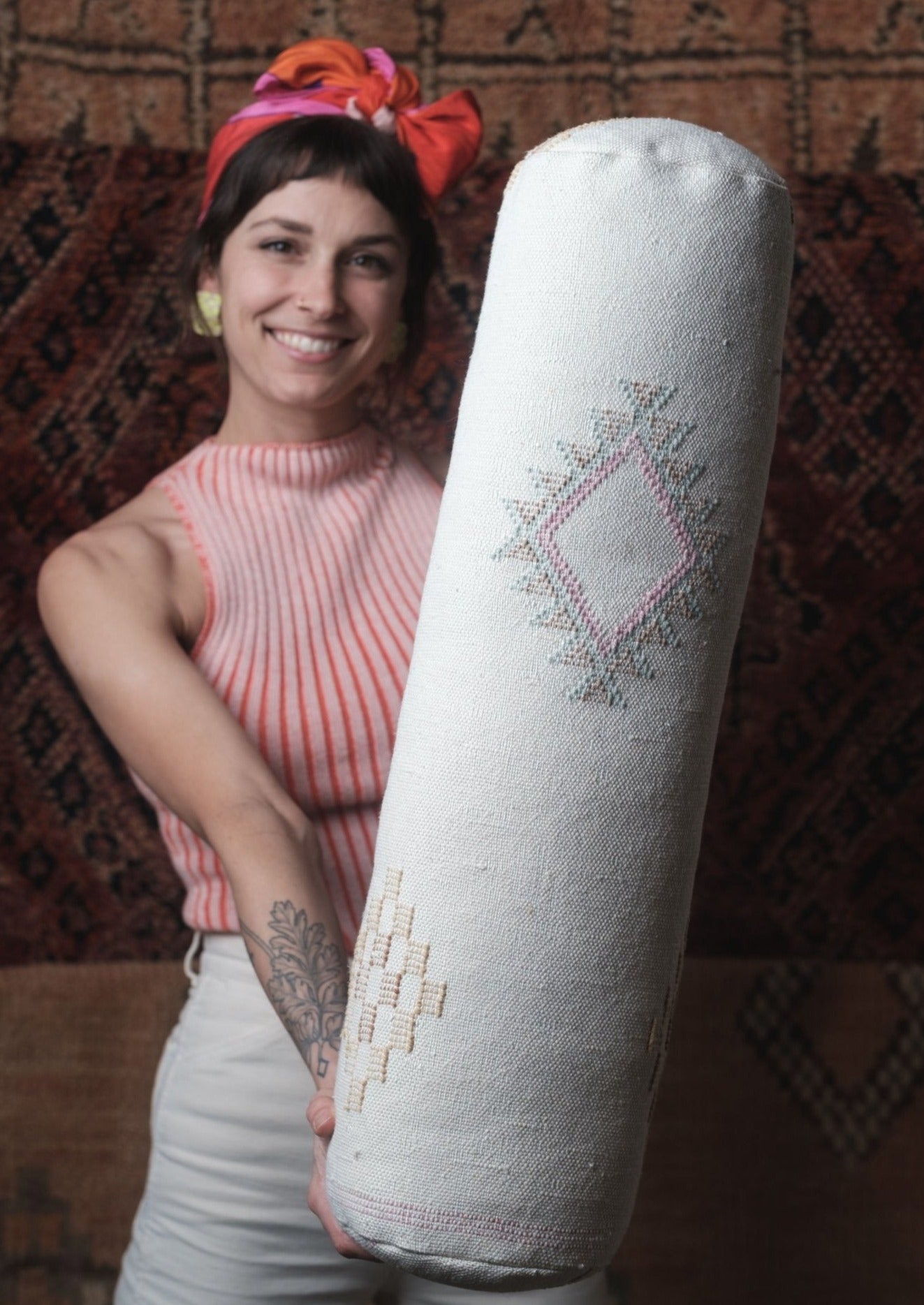 white lumbar pillow made from vegan friendly material with berber symbols hand stitched by artisans of morocco. pretty girl wearing soluna pink top made from organic fibers with pink bow and loud ones green checkered earrings with a rug back drop. 