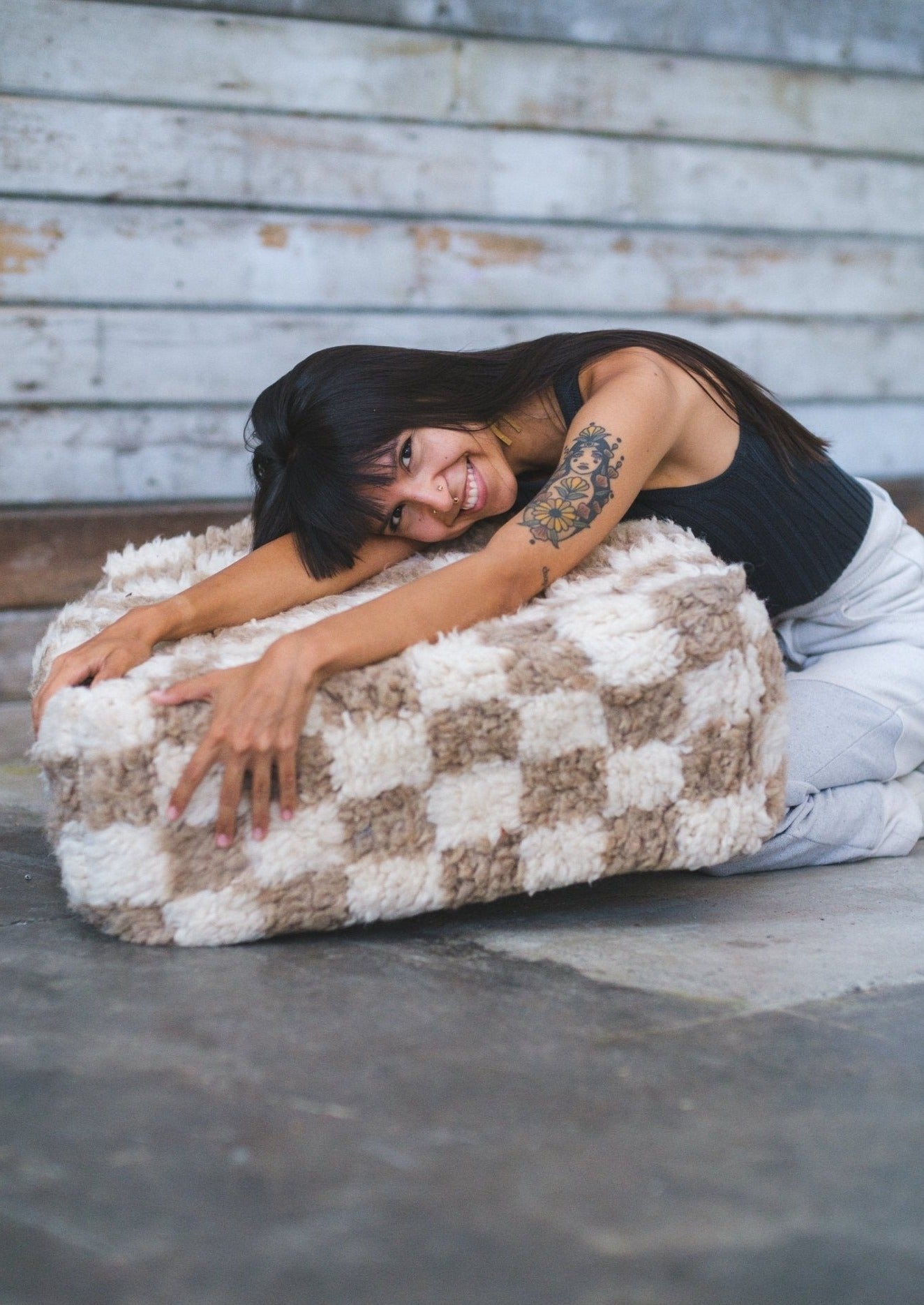 hot girl is beaming a wide smile while holding her favorite moroccan floor cushion made from 100% wool. background wall is rustic and floors are cement. tatoo on left arm bangs and long hair 