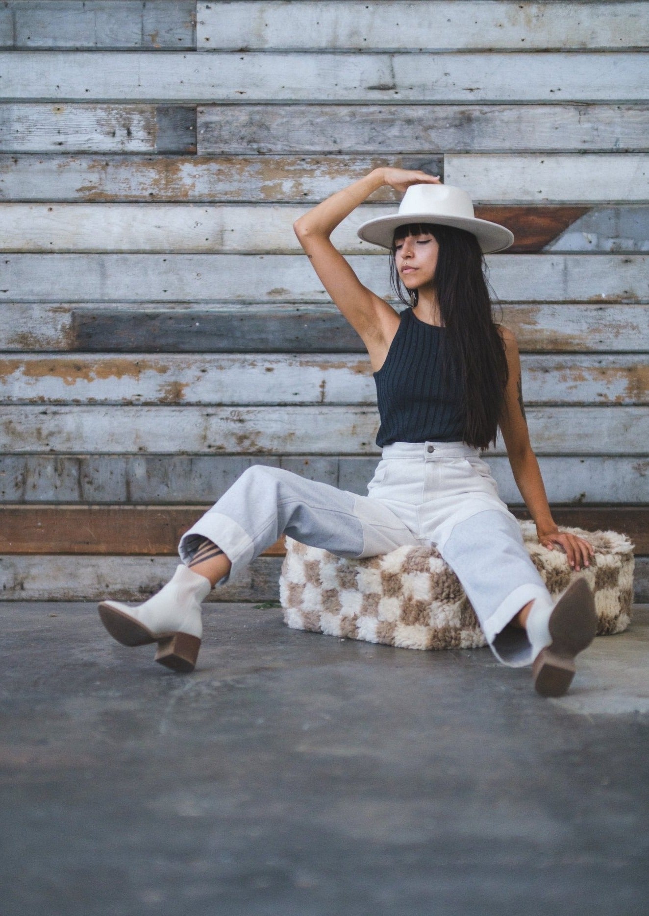girl kick backs and relaxes on a moroccan floor cushion checkered tan and white. she is wearing a white hat and white boots top and bottom are soluna collective made from organic fibers. she is leaning against a rustic wood wall on cement floors
