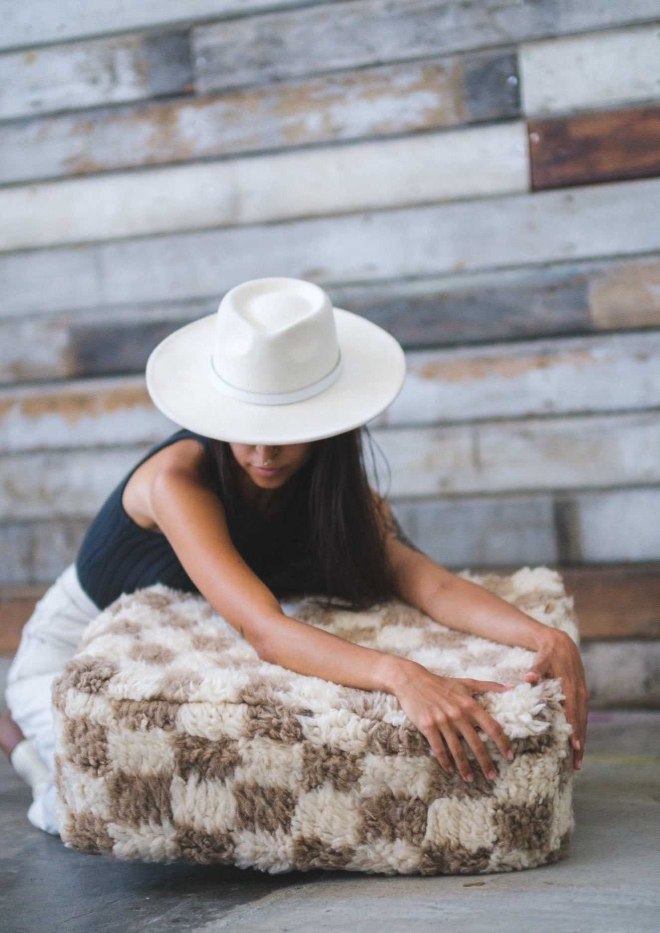 gorgeous and mysterious girl wearing white hat holds and hugs a moroccan floor cushion thats checkered white and tan in a rustic location. 100% wool. 
