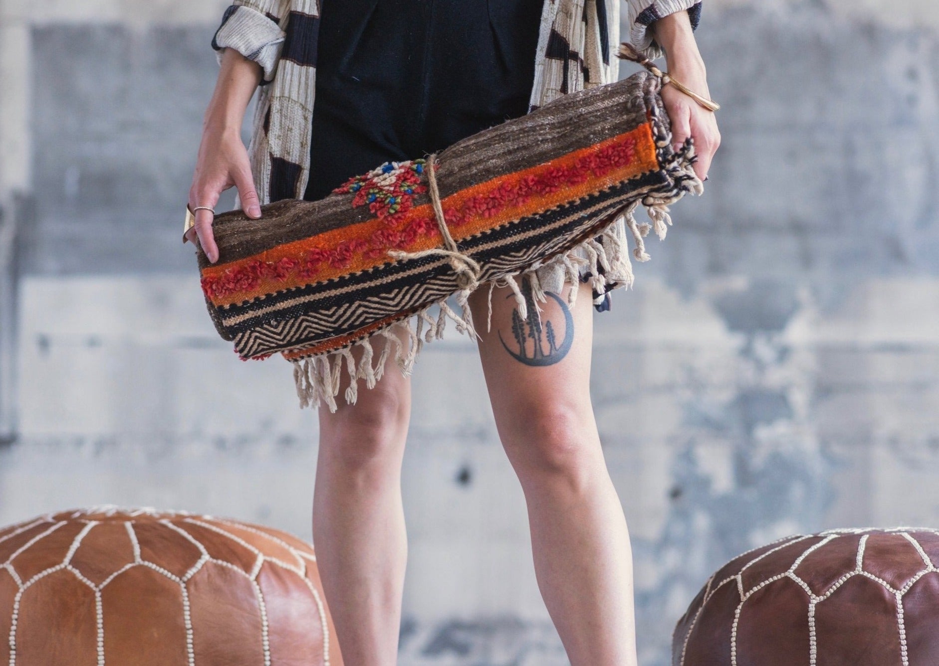 morrocan poufs and textiles go hand in hand. girl holds a handwoven berber textile while standing between a dark pouf and light leather pouf. 