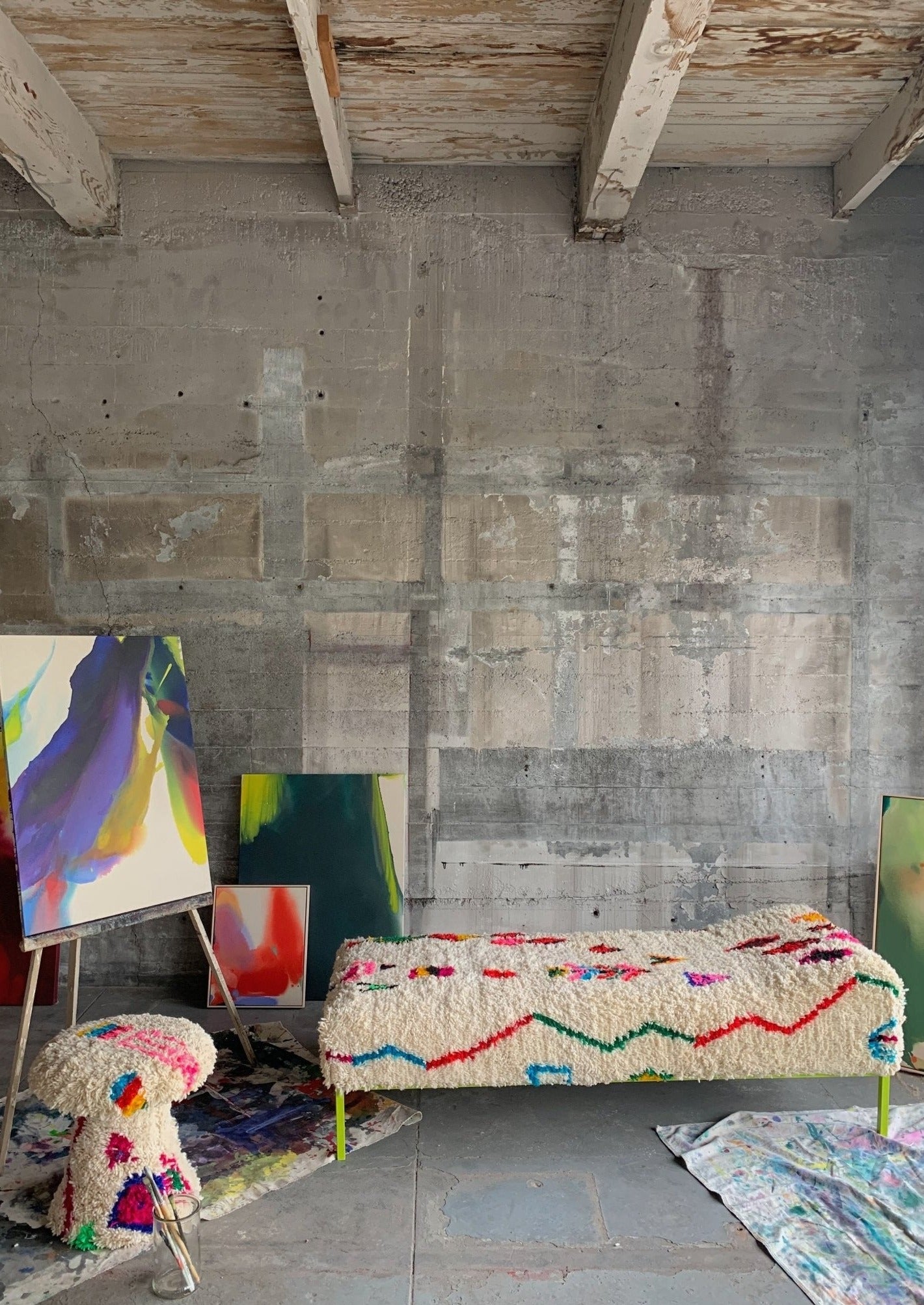 urban rustic art studio scene with mycelia stool and daybed both upholstered with a moroccan rug. Many finished abstract paintings decorate the space. 