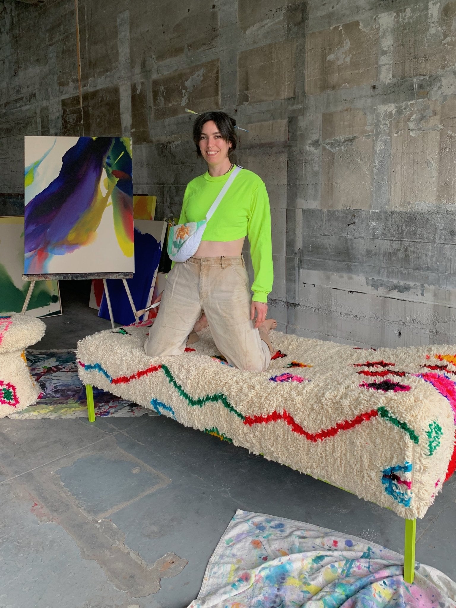 playful portland artists wearing carhart pants and a fanny pack poses by her abstract paintings while kneeling on her upholstered day bed with a moroccan rug. 