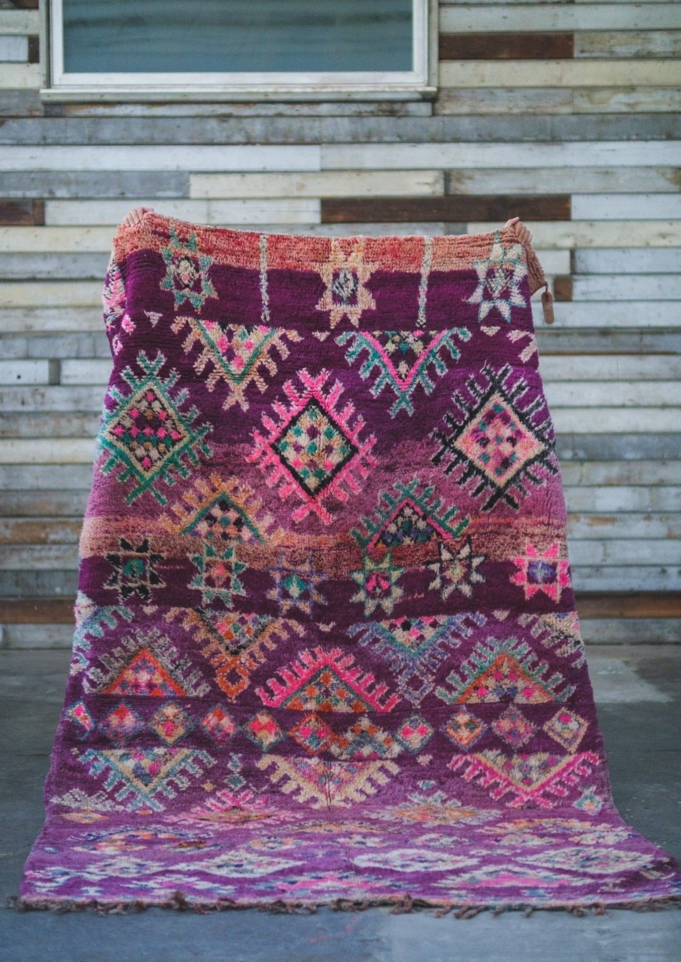playful colors and design decorate this moroccan rug. purple pinks and details are perfect for a girls bedroom. rug is being held up against a rustic wood back drop 