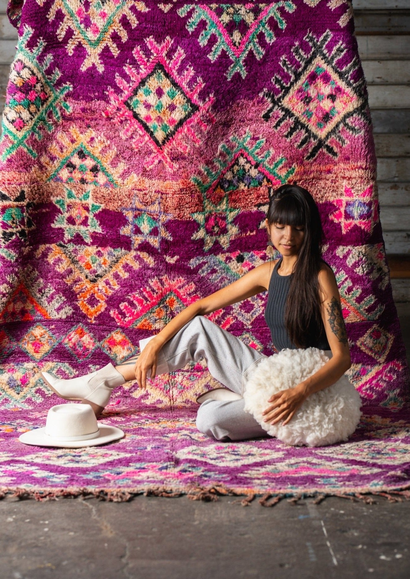 dramatic studio light shows off the color in the rug and highlights the berber motifs. girl sits relaxed on rug holding a sheeps wool round pillow. 