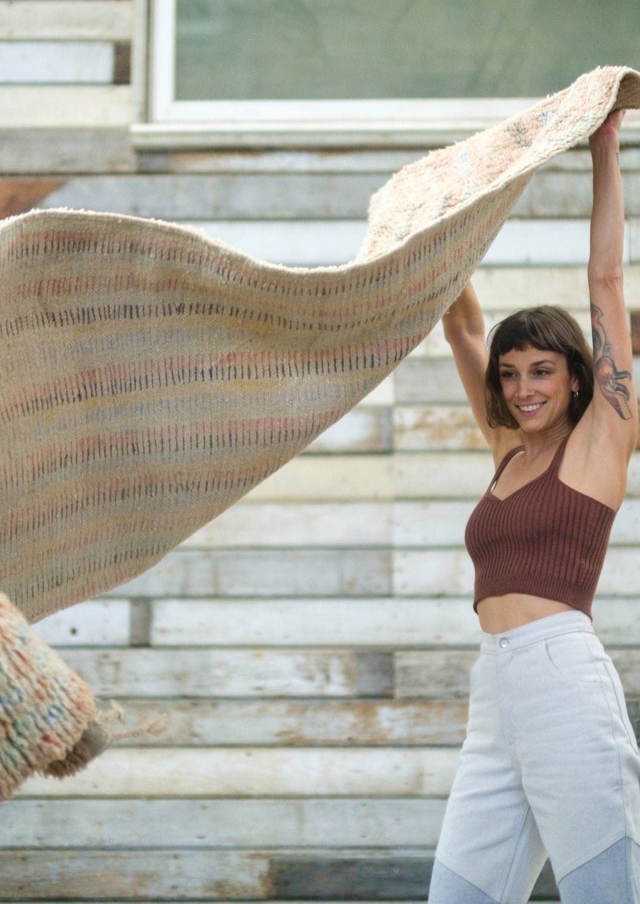 flying carpet feels as this girl rolls out her favorite runner woven by berber women. She is happy with short brown hair and bangs wearing soluna collective pants and top made from organic fibers. She is in a rustic boho barn with a tatoo on her left arm of a fox in motion 