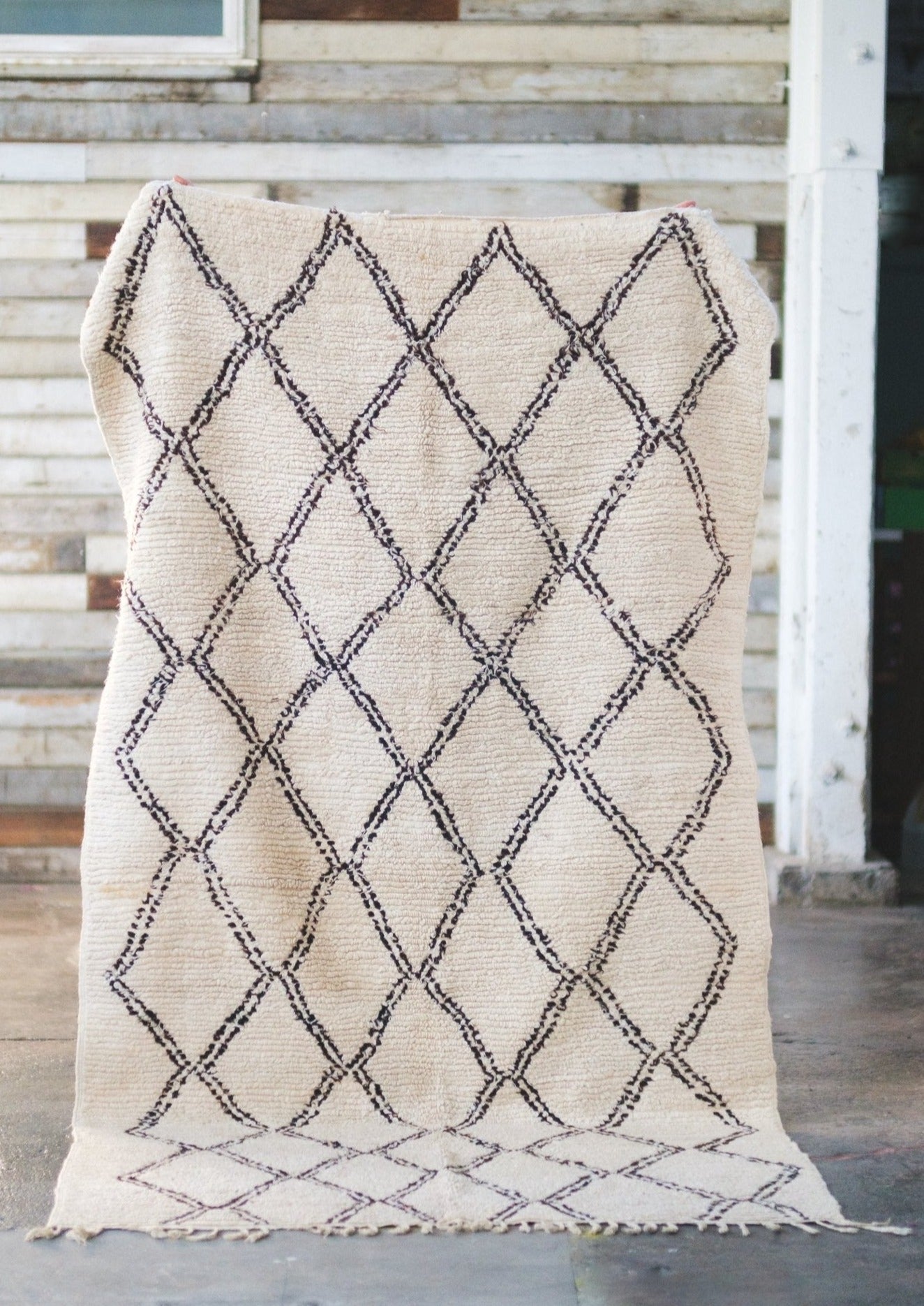 classic beni ourain rug is displayed against a rustic barn wall and cement floors. white wool decorated with black diamonds all natural dye 