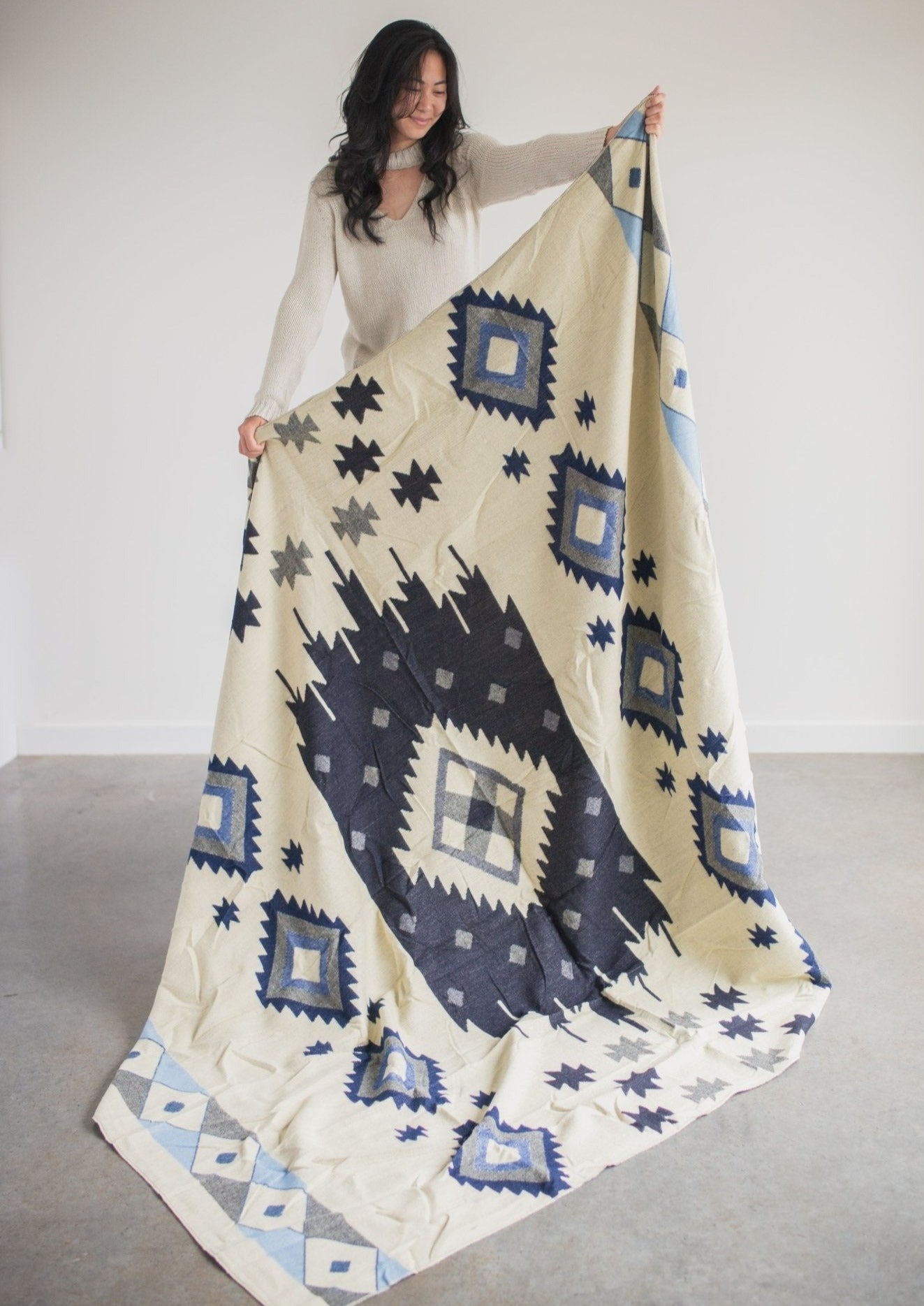asian girl wearing soft sweater holds up reverse side of alpaca blanket so you can see the full design. 