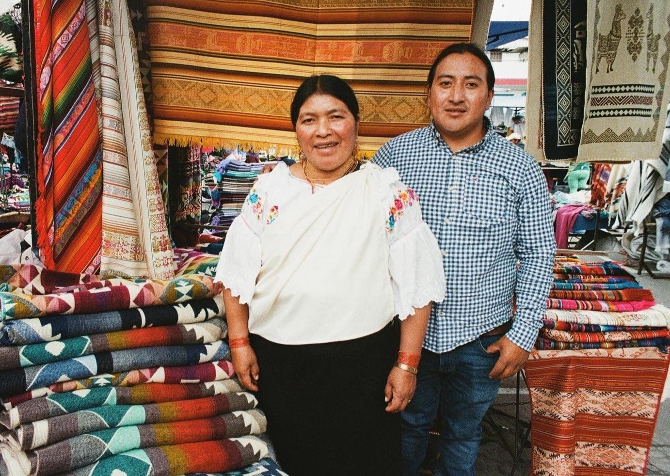 artisans of Ecuador stand outside at their marketplace by the handwoven textiles they sell.