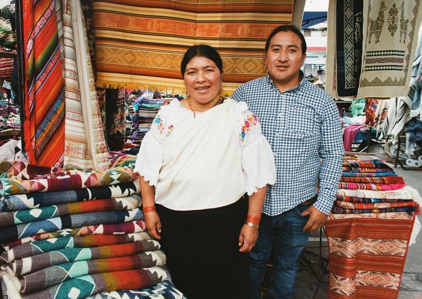 Artisans of Ecuador stand outside at a market place in Otavalo by their handmade textiles 