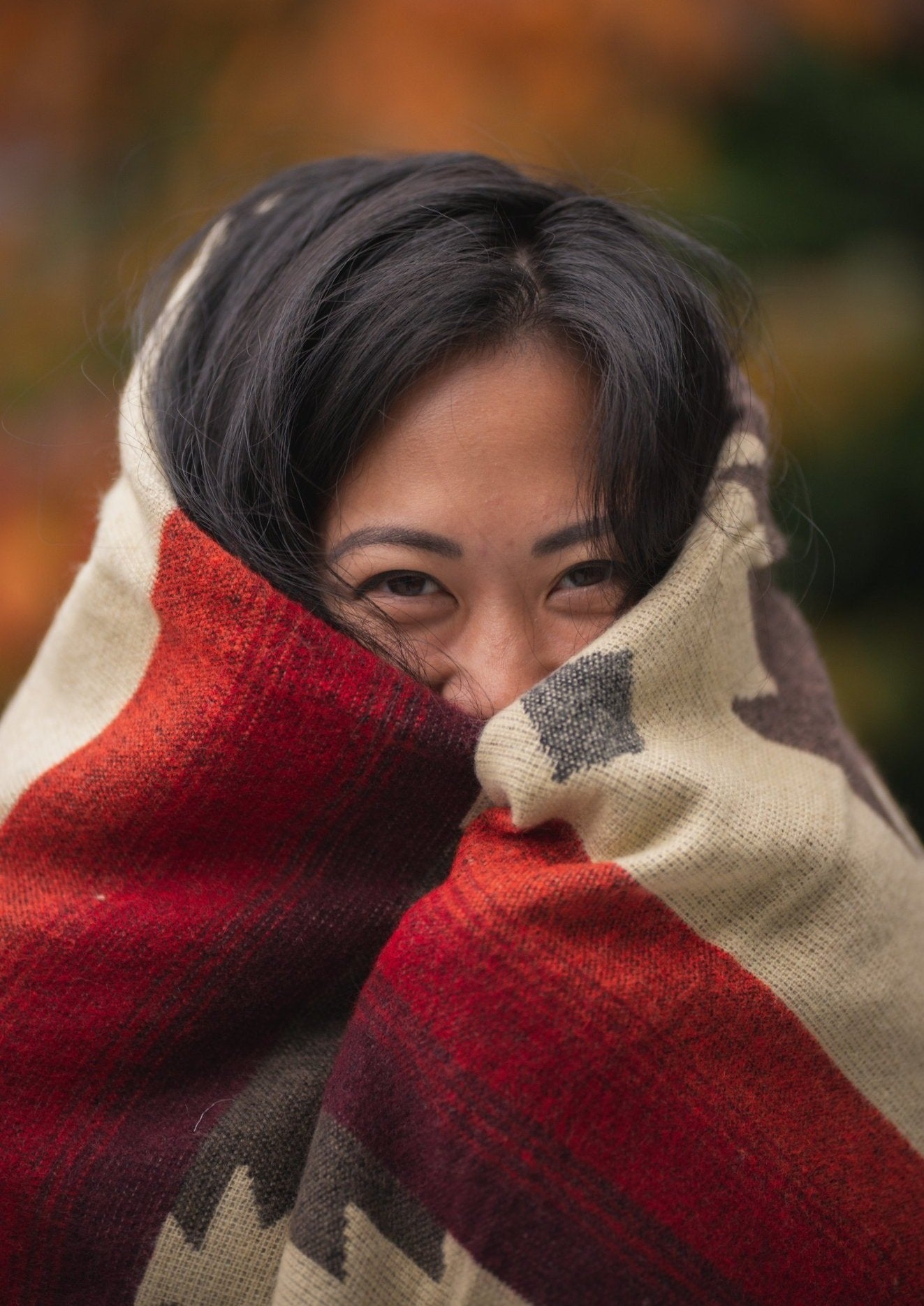 Asian girl wraps up in a cozy alpaca blanket outside during fall. The blanket keeps her face warm and her eyes are smiling. 